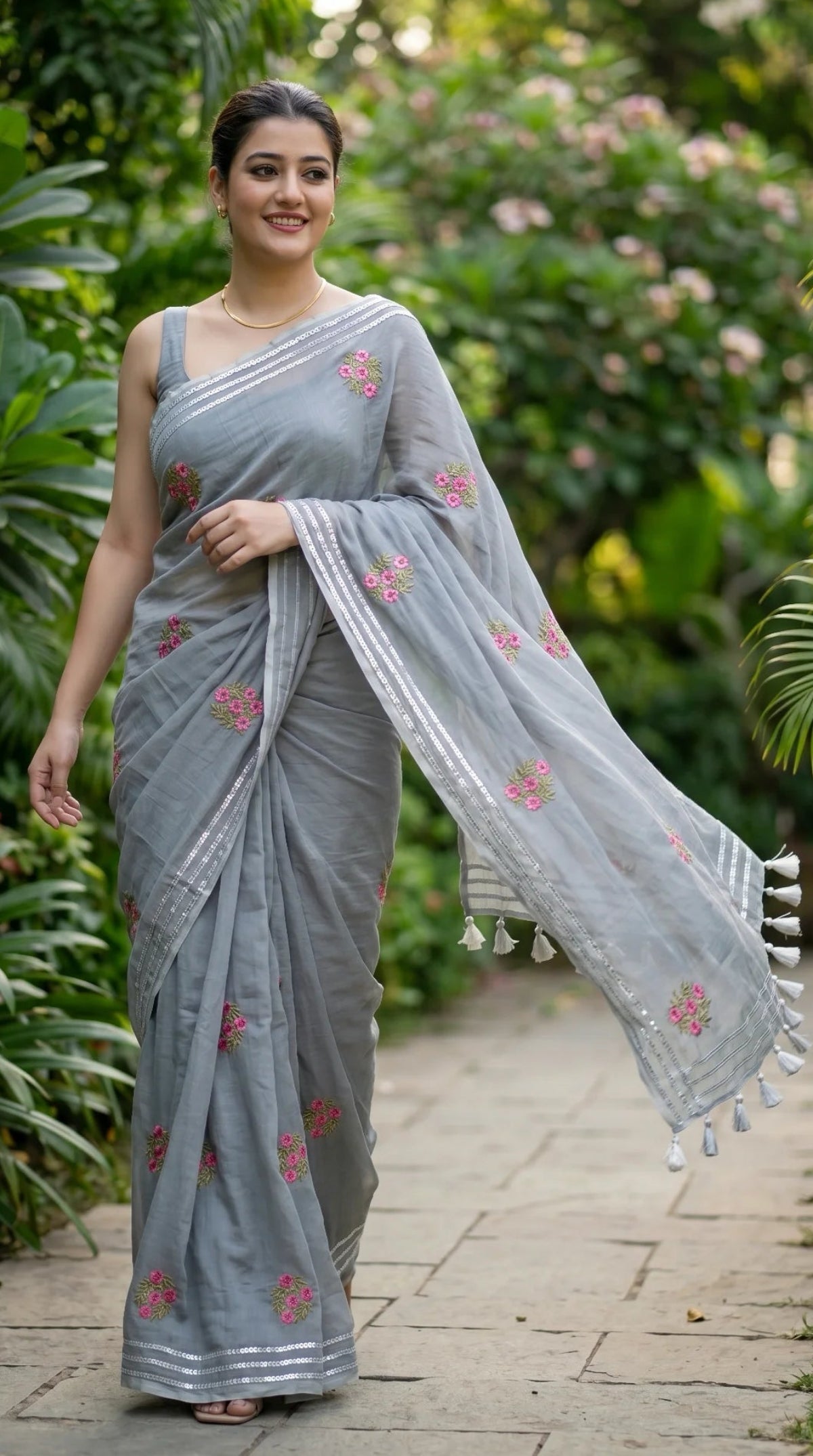 Woman in a light blue saree with floral patterns standing outdoors among greenery.