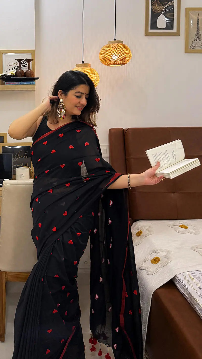 Woman in a black soft saree with red heart embroidery in a living room setting.