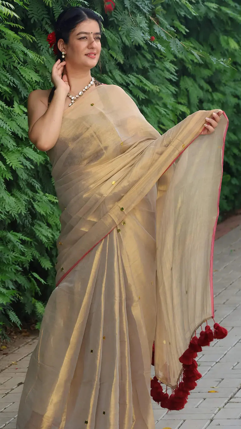 Woman in a hilary gold tissue saree with red accents standing outdoors against green foliage.