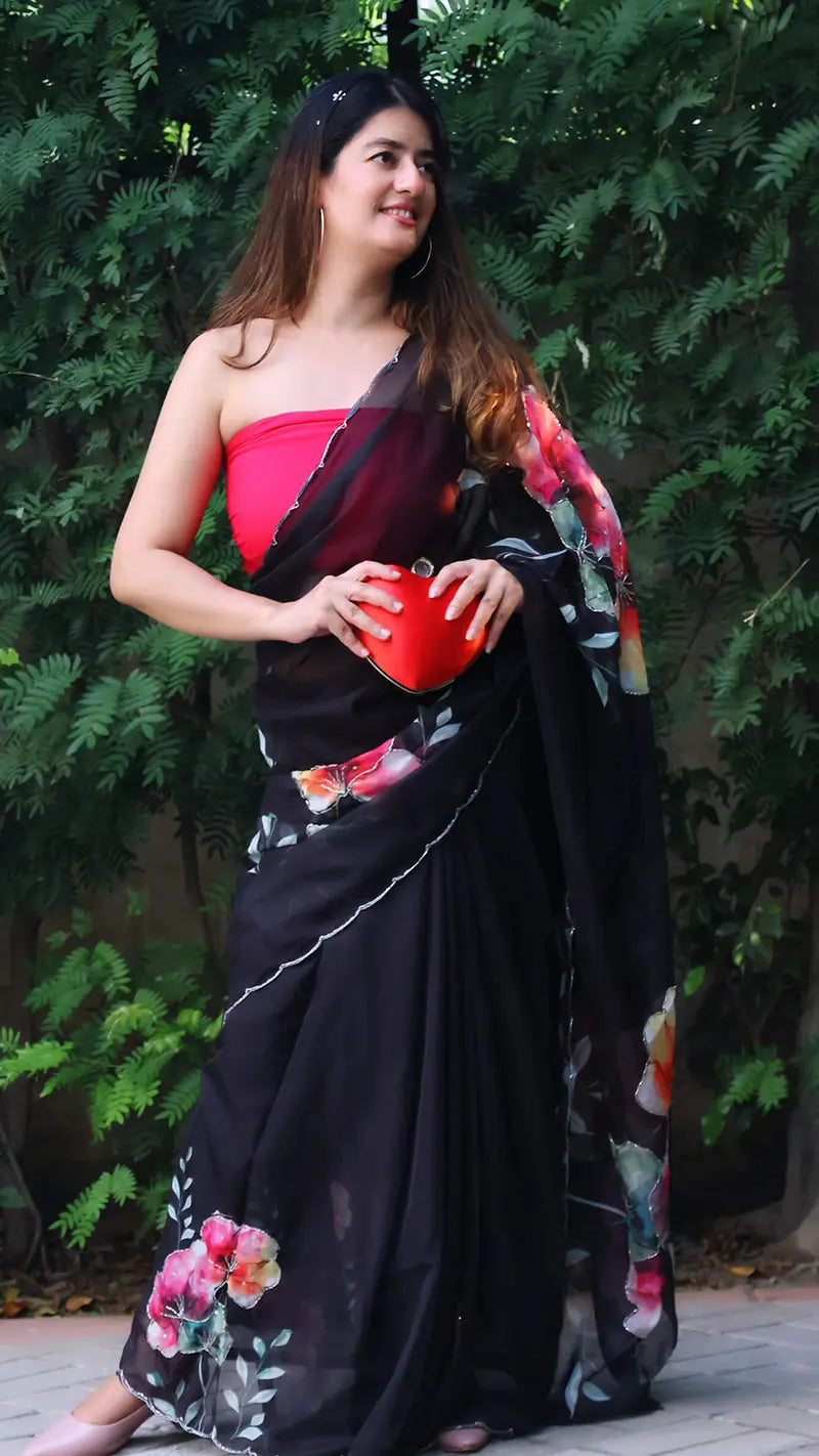 Woman in a black organza saree with floral patterns holding a red heart-shaped object, standing against a green leafy background.