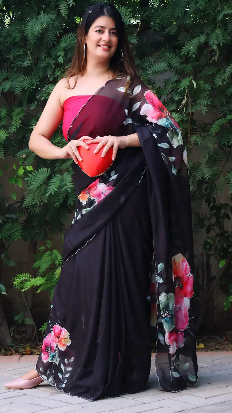 Woman in a black saree with floral patterns holding a red clutch against a green foliage background