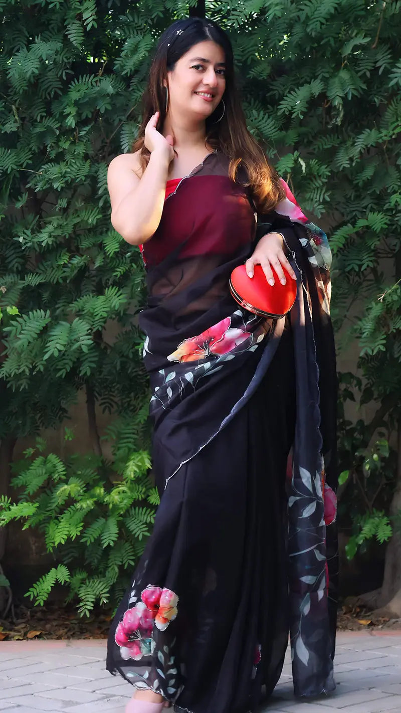 Woman in a black saree with floral patterns holding a red clutch against a green foliage background