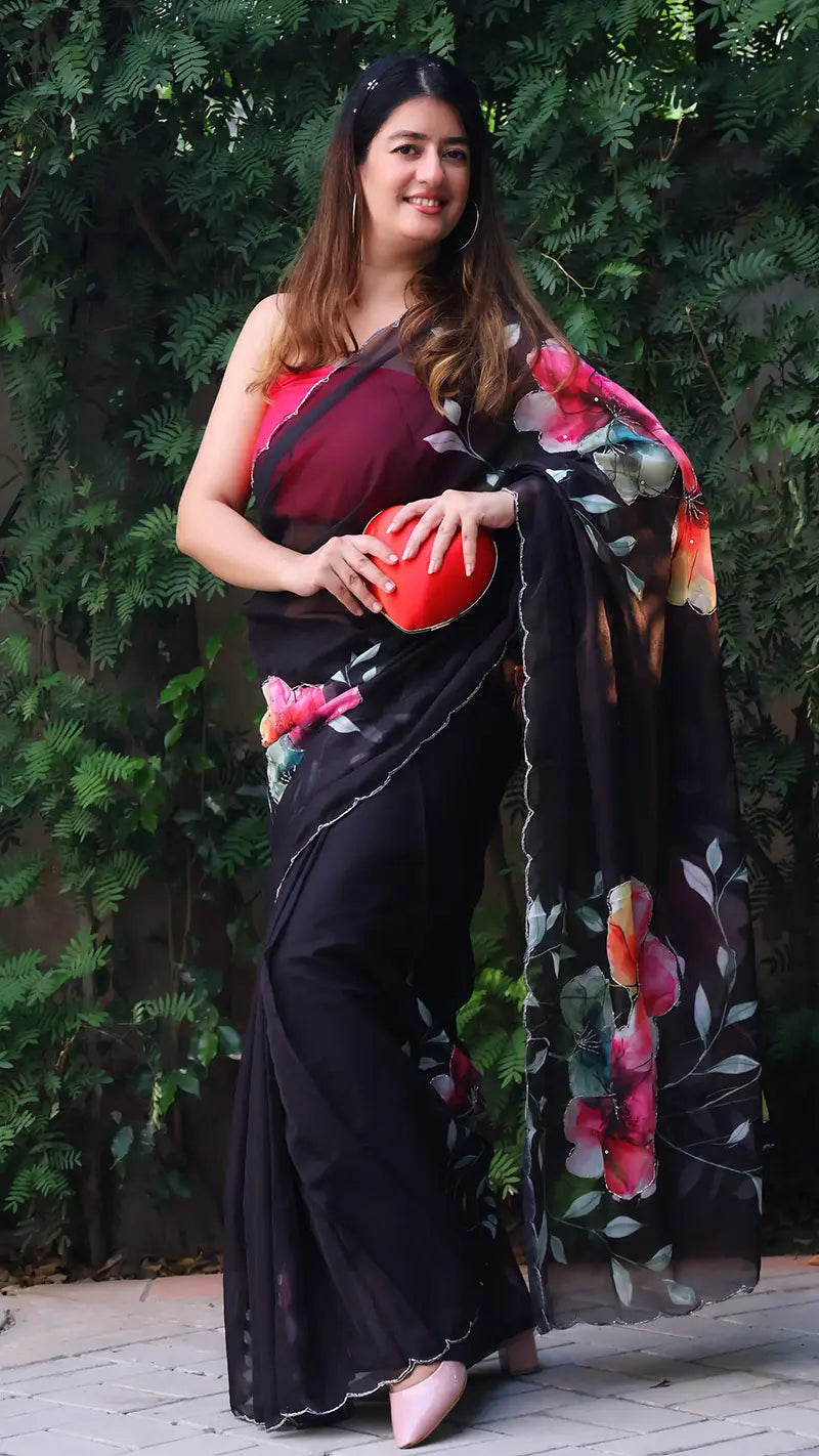 Woman wearing a black organza saree with floral patterns, standing against a green leafy background.