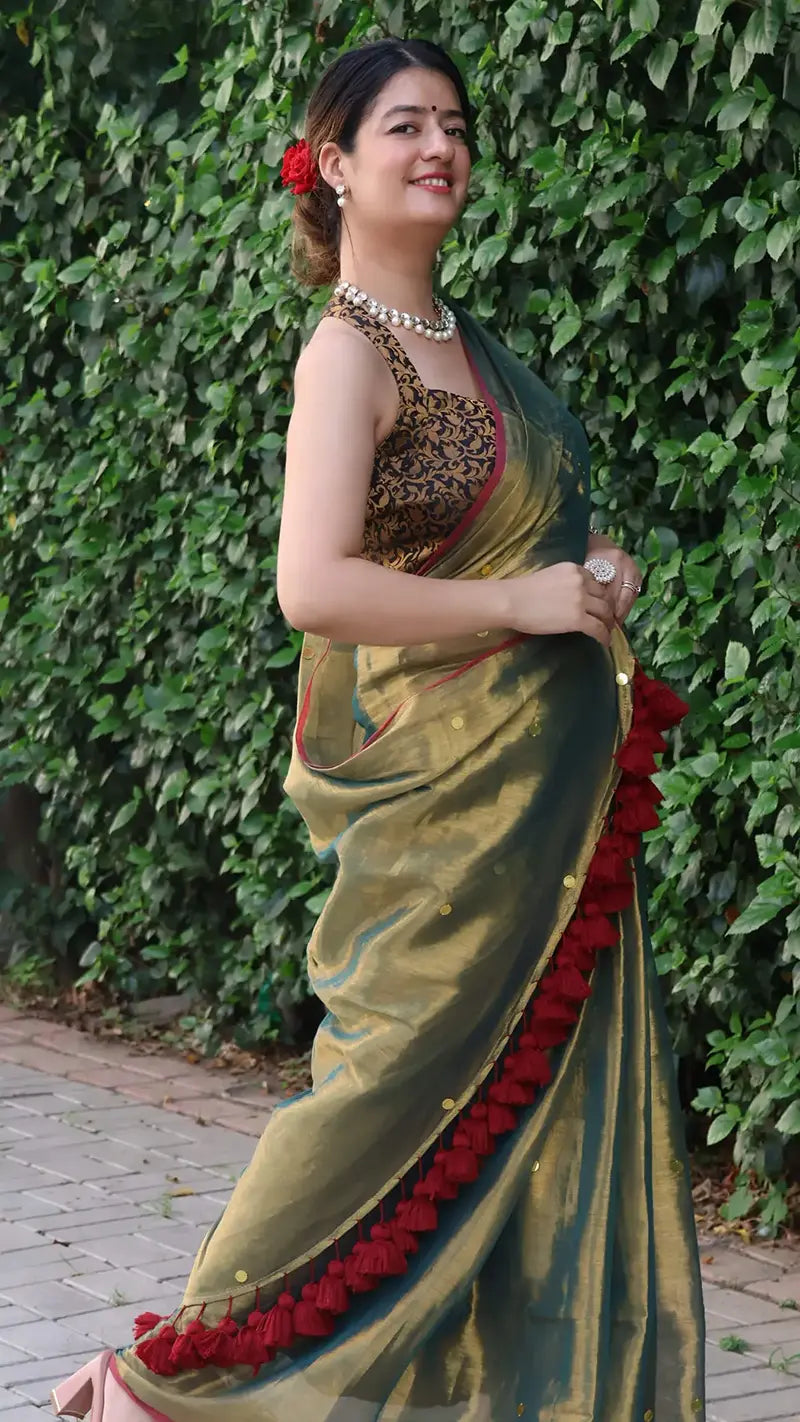 Woman in a Museli Gold Elegant Tissue Saree with a red border standing against a green leafy background