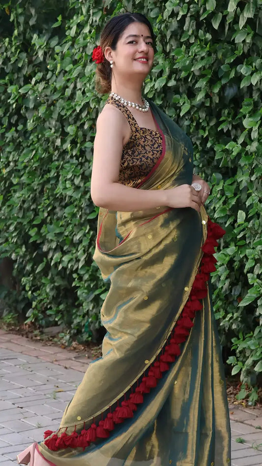 Woman in a Museli Gold Elegant Tissue Saree with a red border standing against a green leafy background