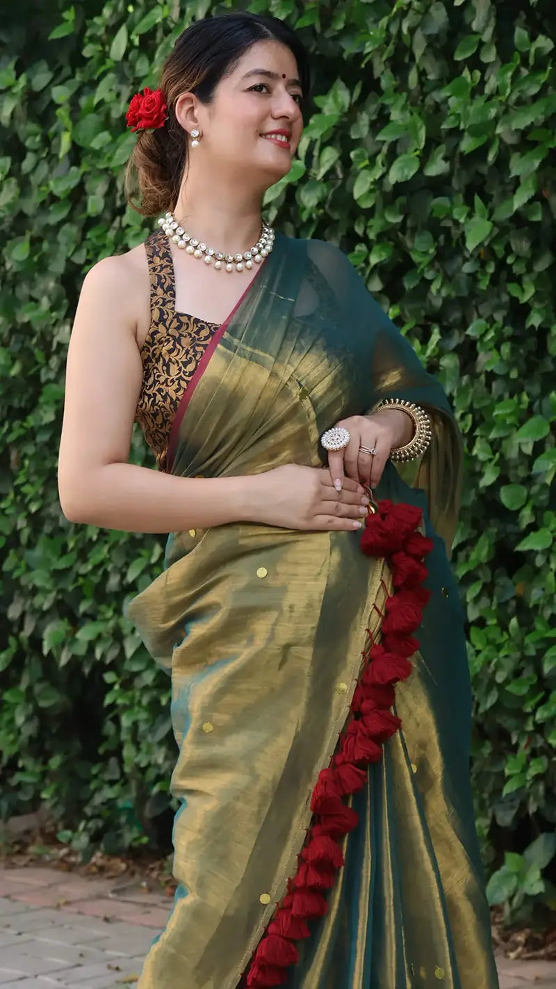 Woman in a Museli Gold Demure Elegant Tissue Saree with red tassels against a green leafy background