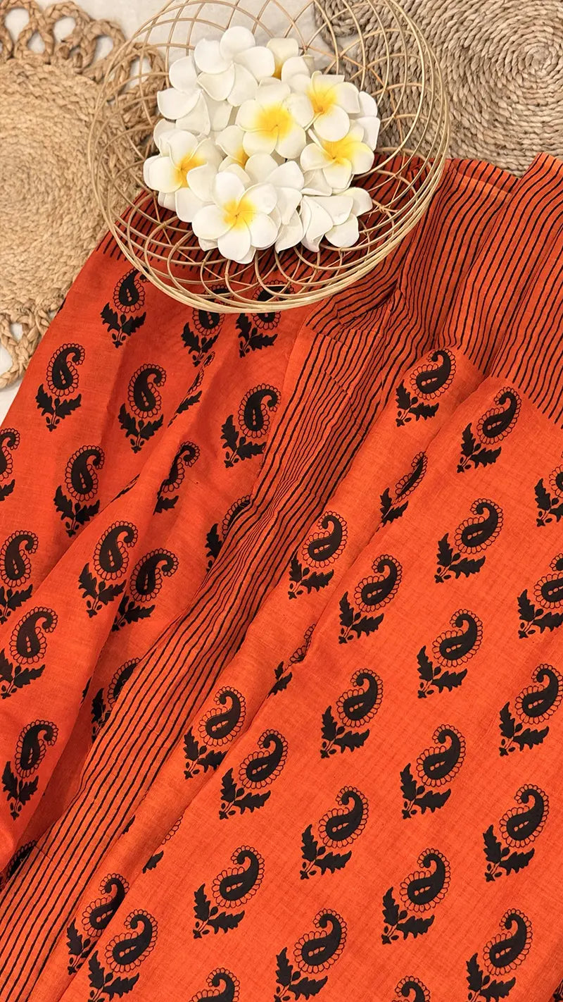Orange saree with black paisley patterns and a small basket of white flowers on a textured surface.