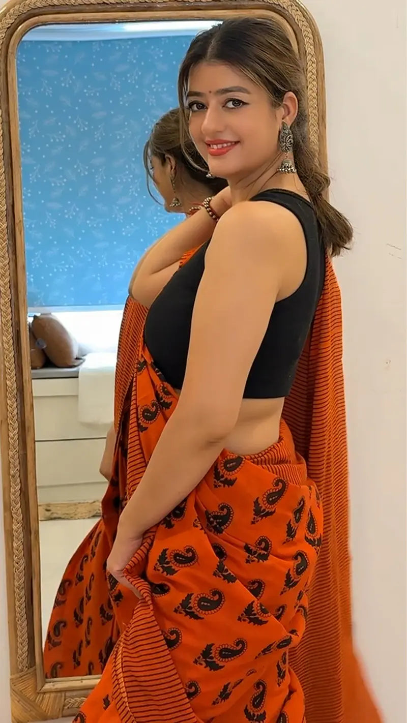 Woman in an orange saree with a black blouse standing in front of a mirror.