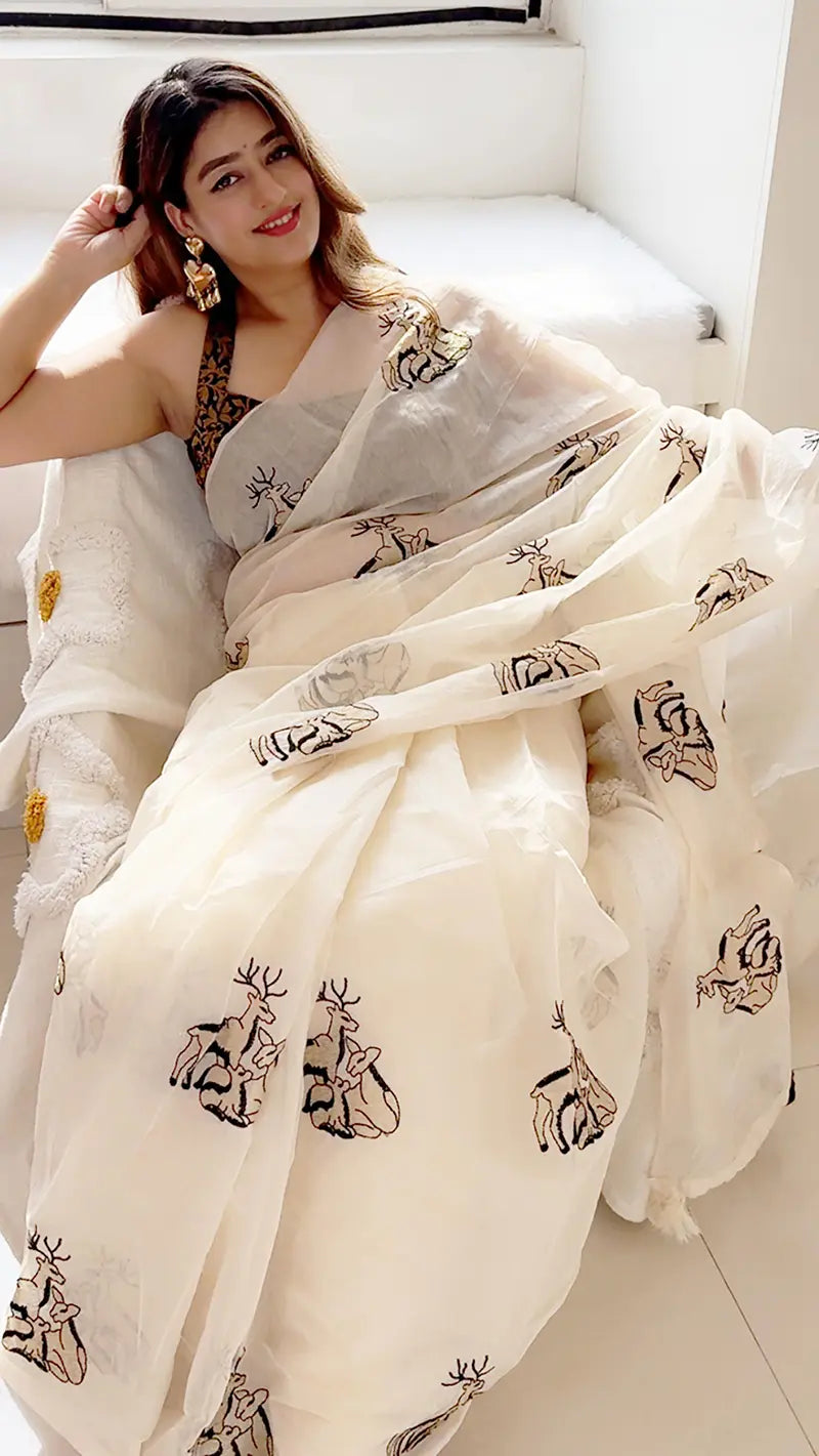 Woman wearing a white saree with black patterns, sitting on a white couch.