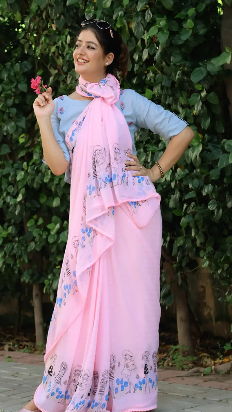 Woman wearing a light pink chiffon saree with a floral pattern, standing outdoors against a green leafy background.