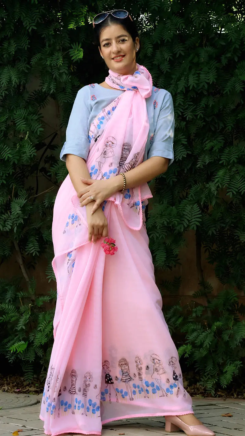 Woman wearing a pink chiffon saree with a floral design, standing against a green leafy background.