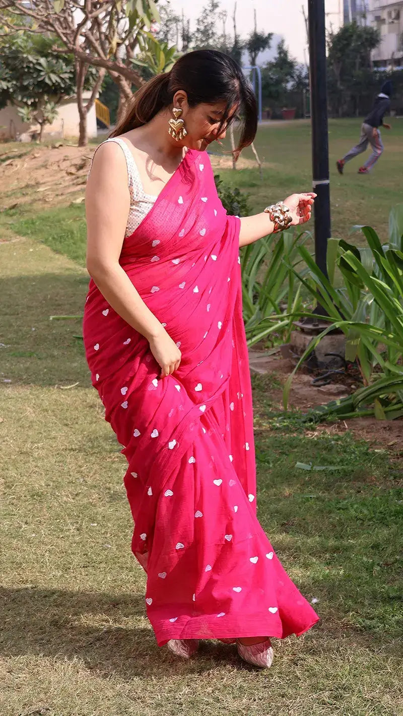 Woman in a bright pink mul cotton saree with white heart embroidery, standing outdoors on grass.