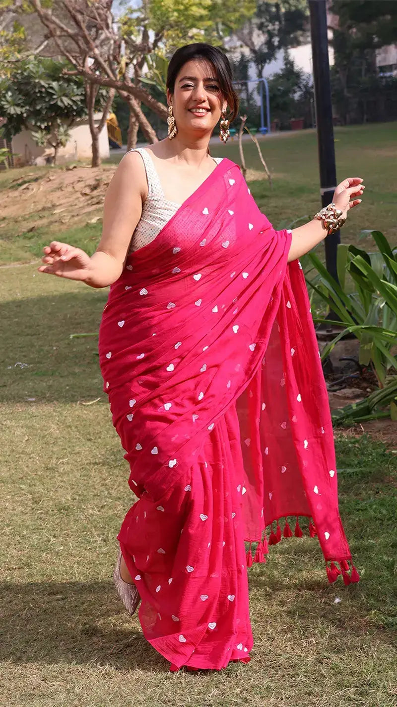Woman in a bright pink mul cotton saree with white heart embroidery standing outdoors on grass.