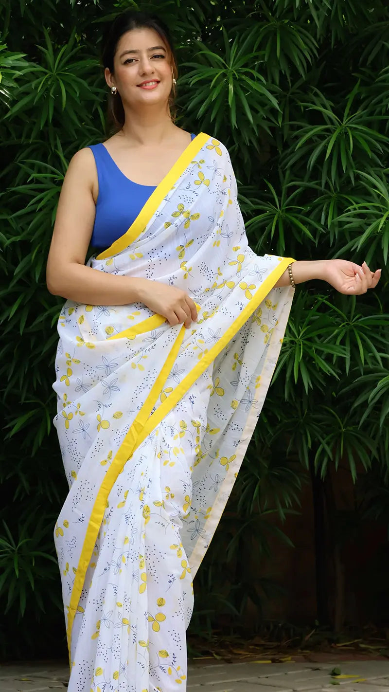 Woman wearing a white linen saree with yellow border and blue blouse against a green leafy background
