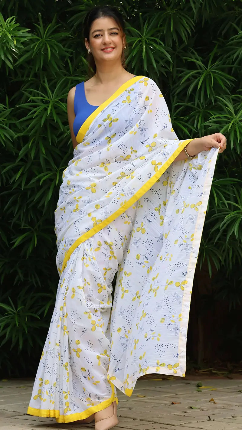 Woman wearing a white linen saree with yellow patterns and a blue blouse, standing against a green leafy background.