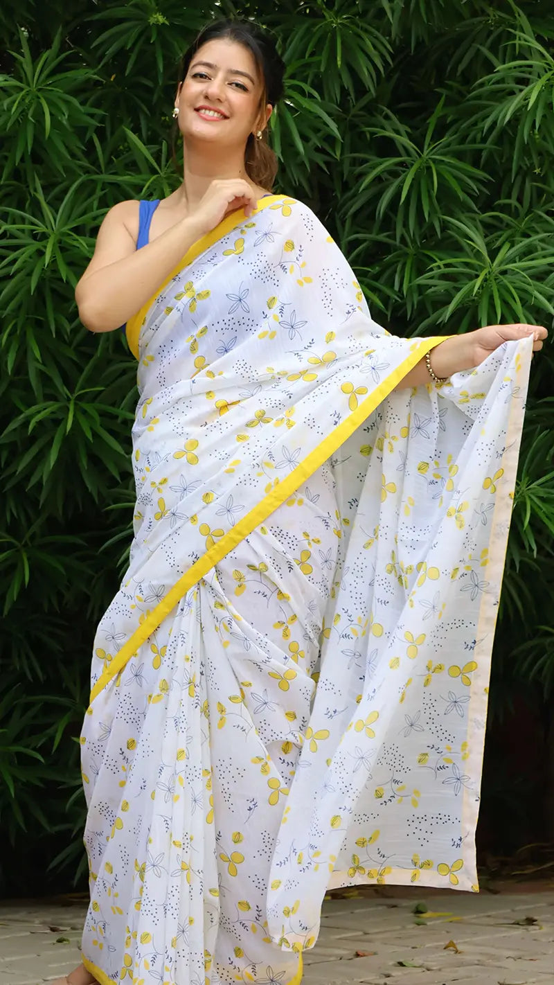 Woman wearing a white  linen saree with yellow patterns and border, standing against a green leafy background.