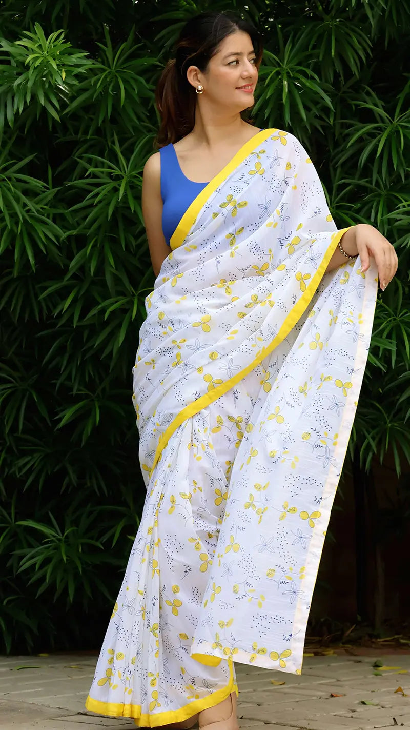 Woman wearing a white linen saree with yellow patterns and border against a green leafy background