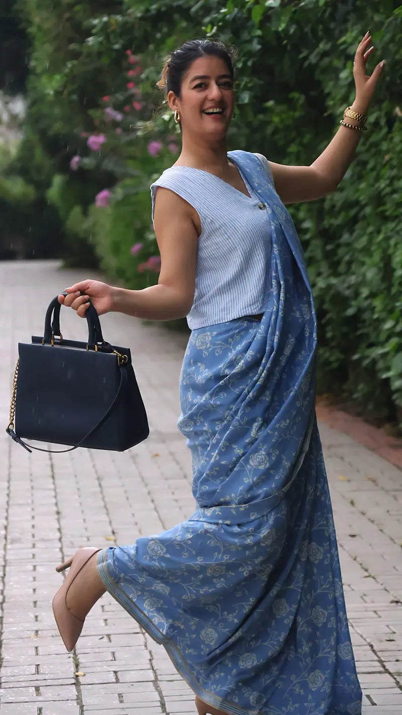 Woman in a blue muslin print saree with a black handbag, standing outdoors with greenery in the background.