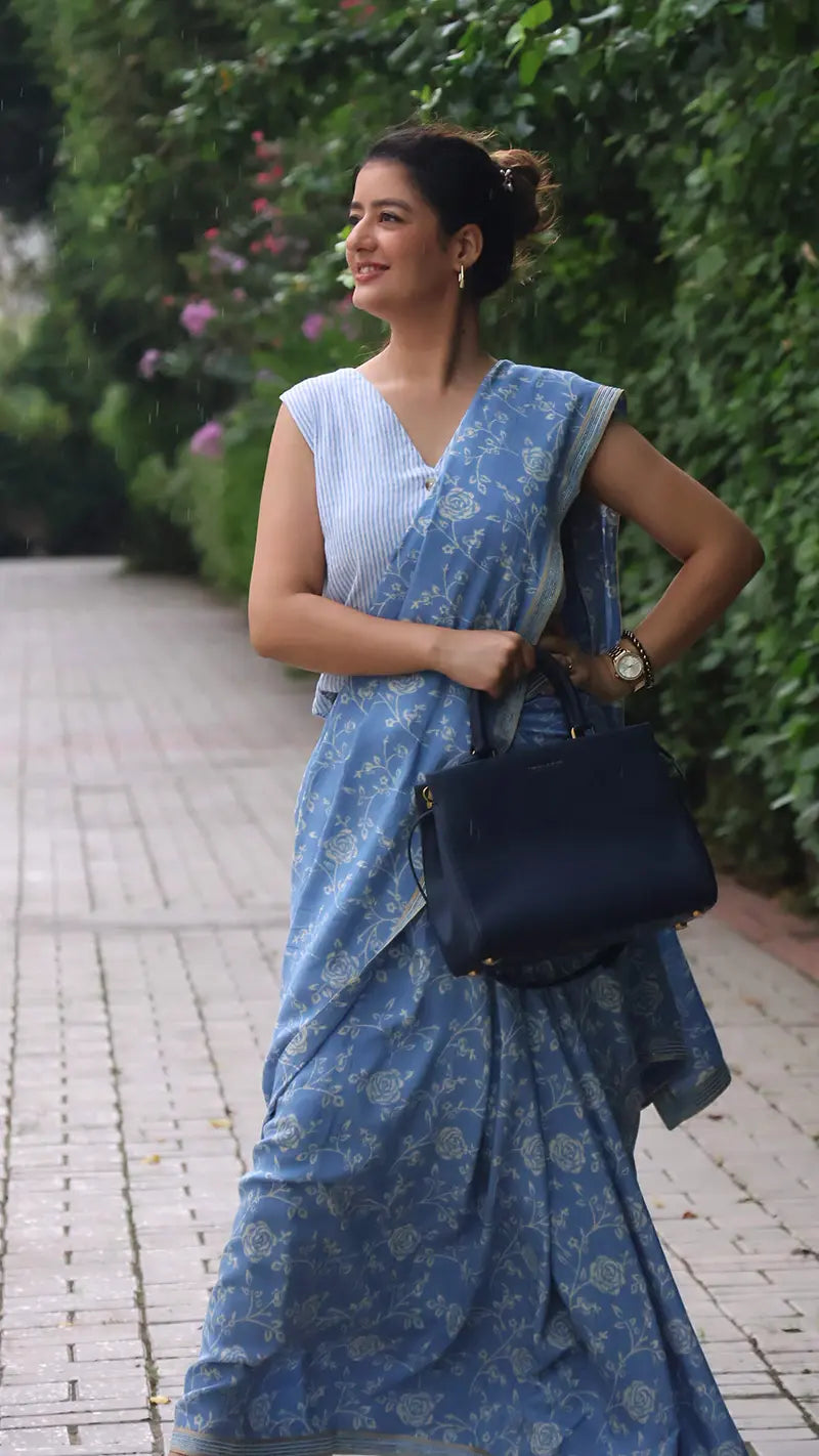 Woman in a blue muslin print saree with a black handbag standing outdoors.
