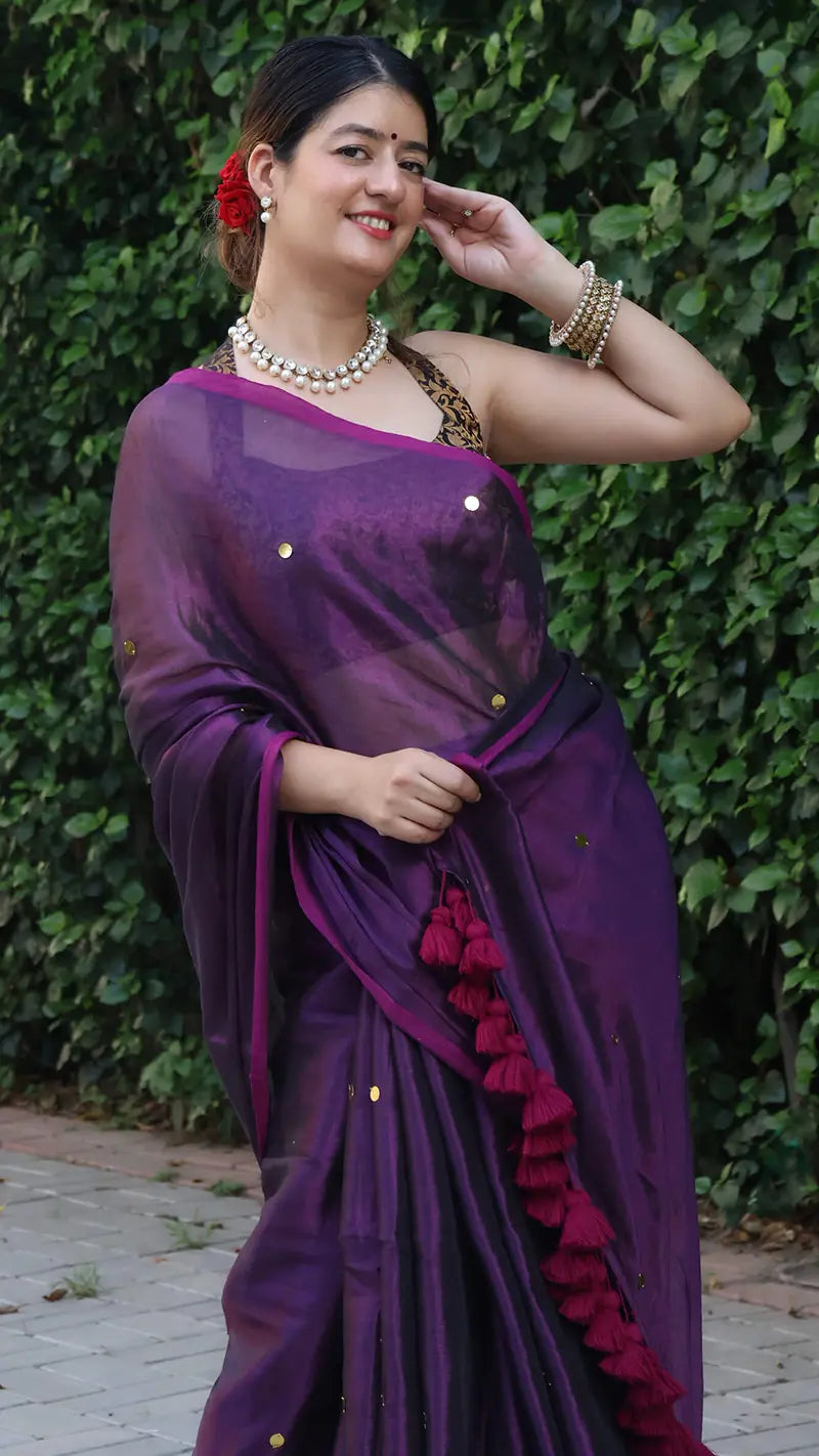 Woman in a purple tissue saree with red details against a green hedge background