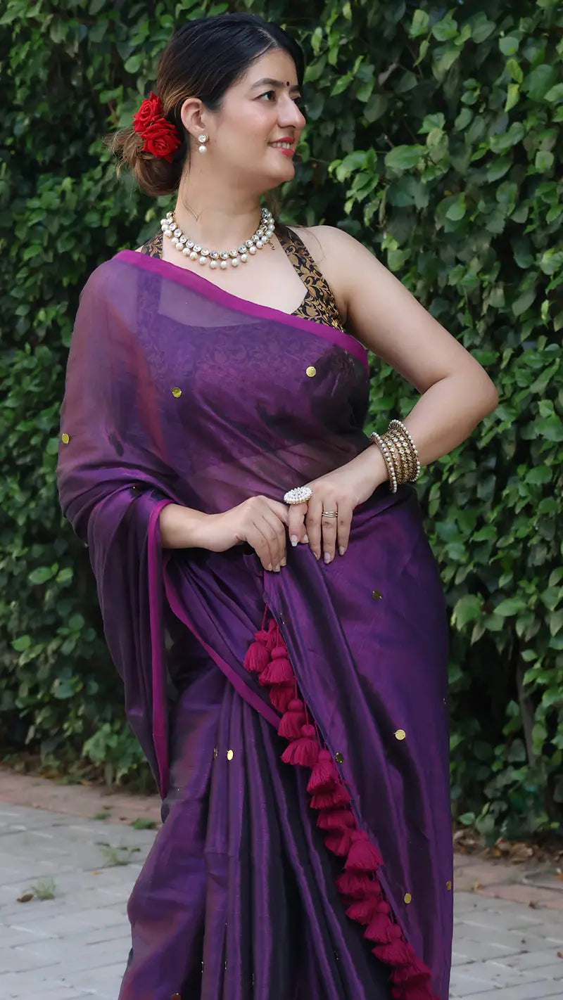 Woman wearing a purple tissue saree with a red flower in her hair against a green leafy background