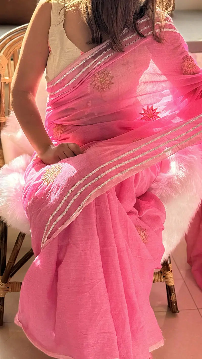 Person wearing a pink saree with floral patterns, sitting on a chair.