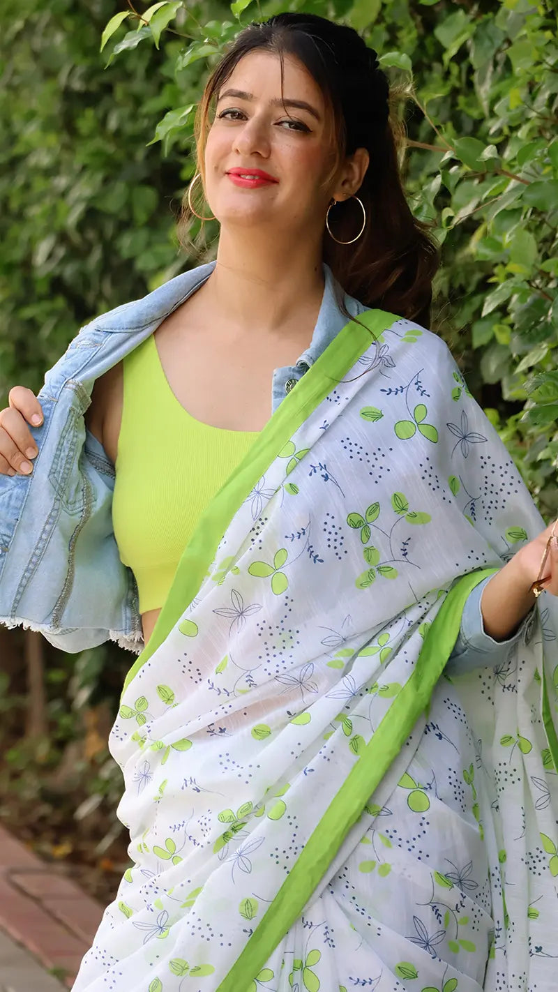 Woman wearing a white saree with green border and green top, standing against a green leafy background.