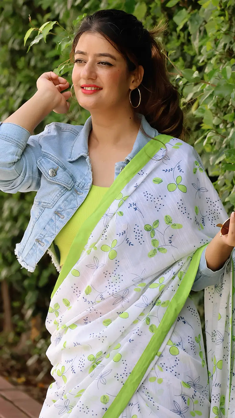 Woman wearing a white saree with green border and denim jacket against a green leafy background