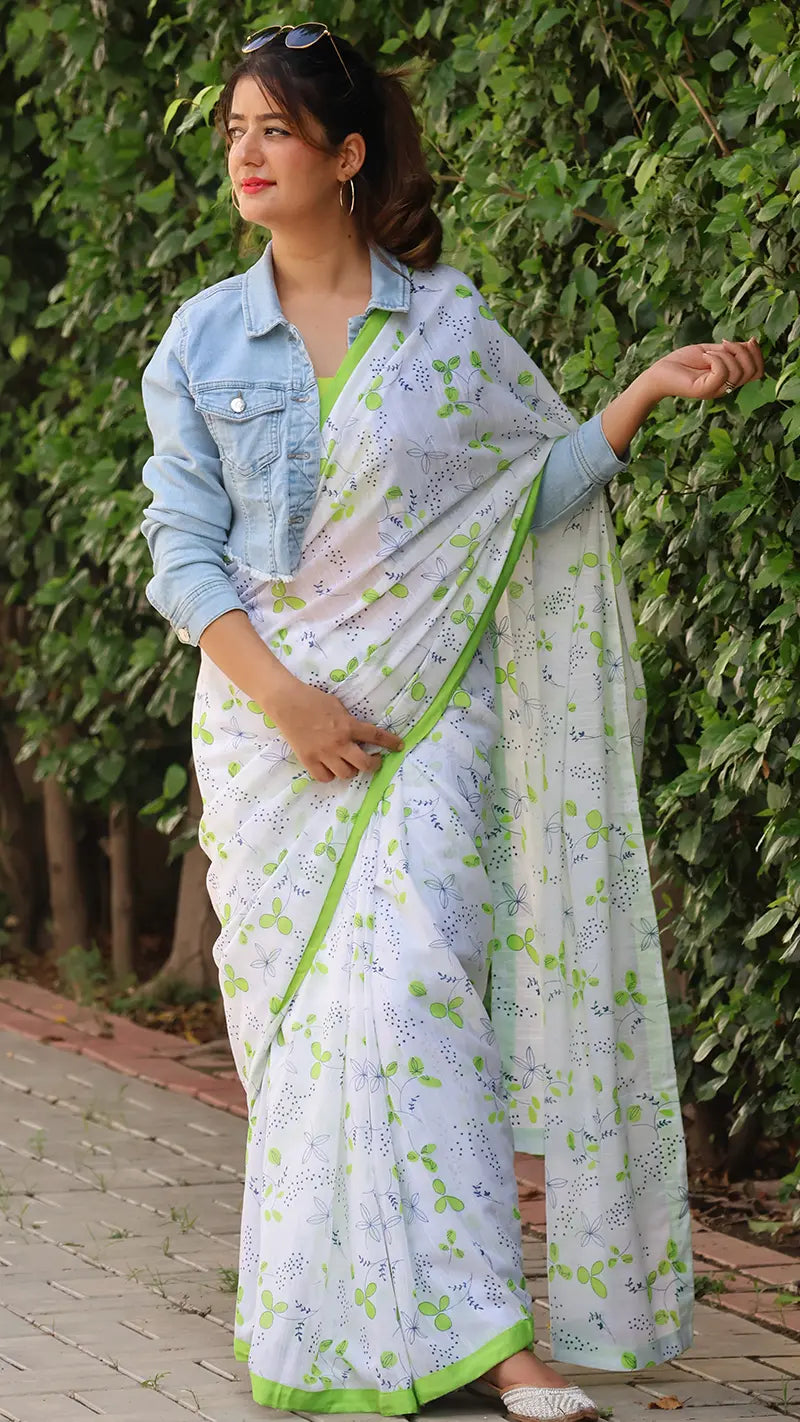 Woman wearing a white linen saree with green border and denim jacket against a green leafy background