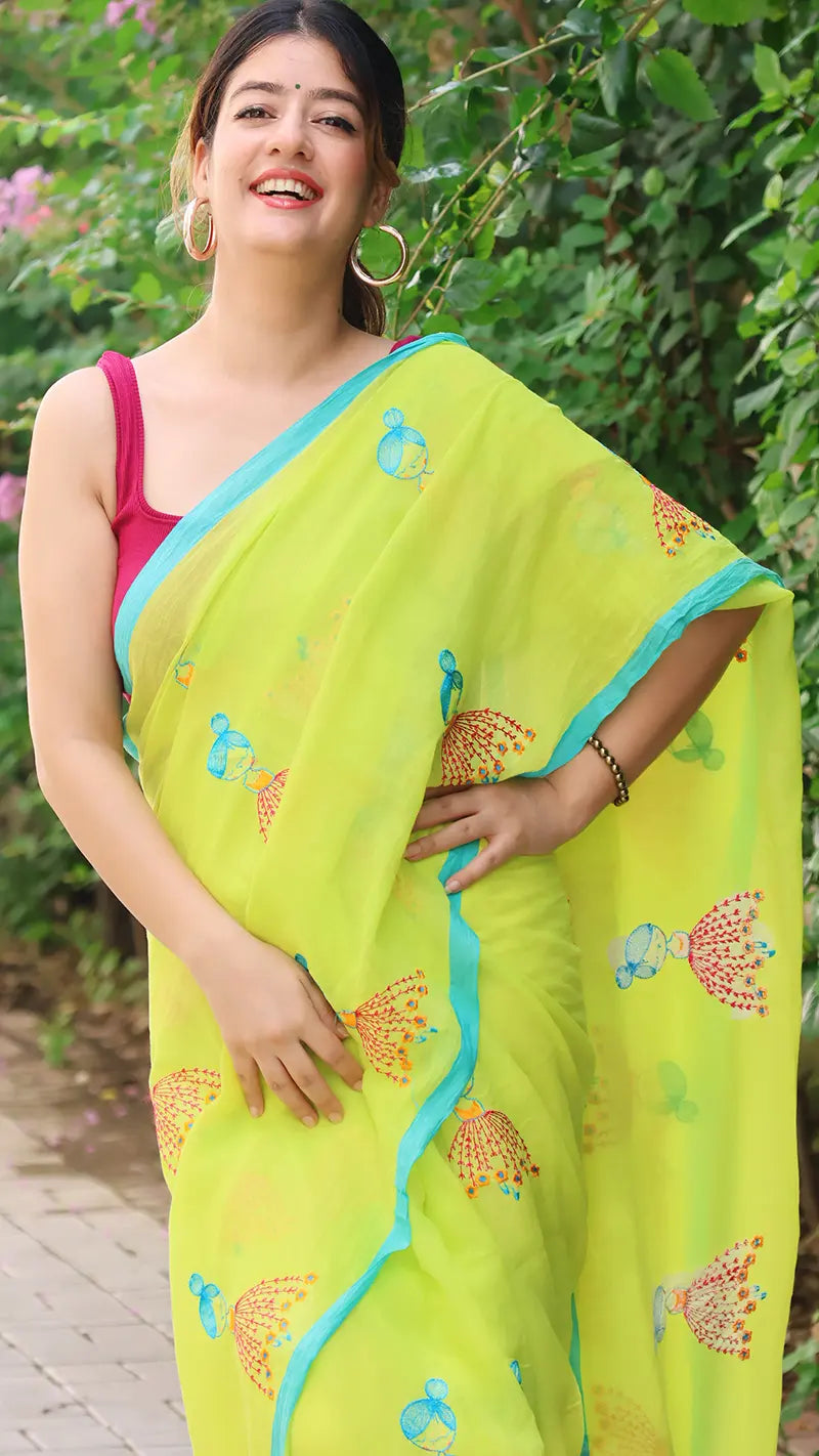 Woman wearing a bright mul cotton saree with a maroon blouse, standing outdoors with greenery in the background.