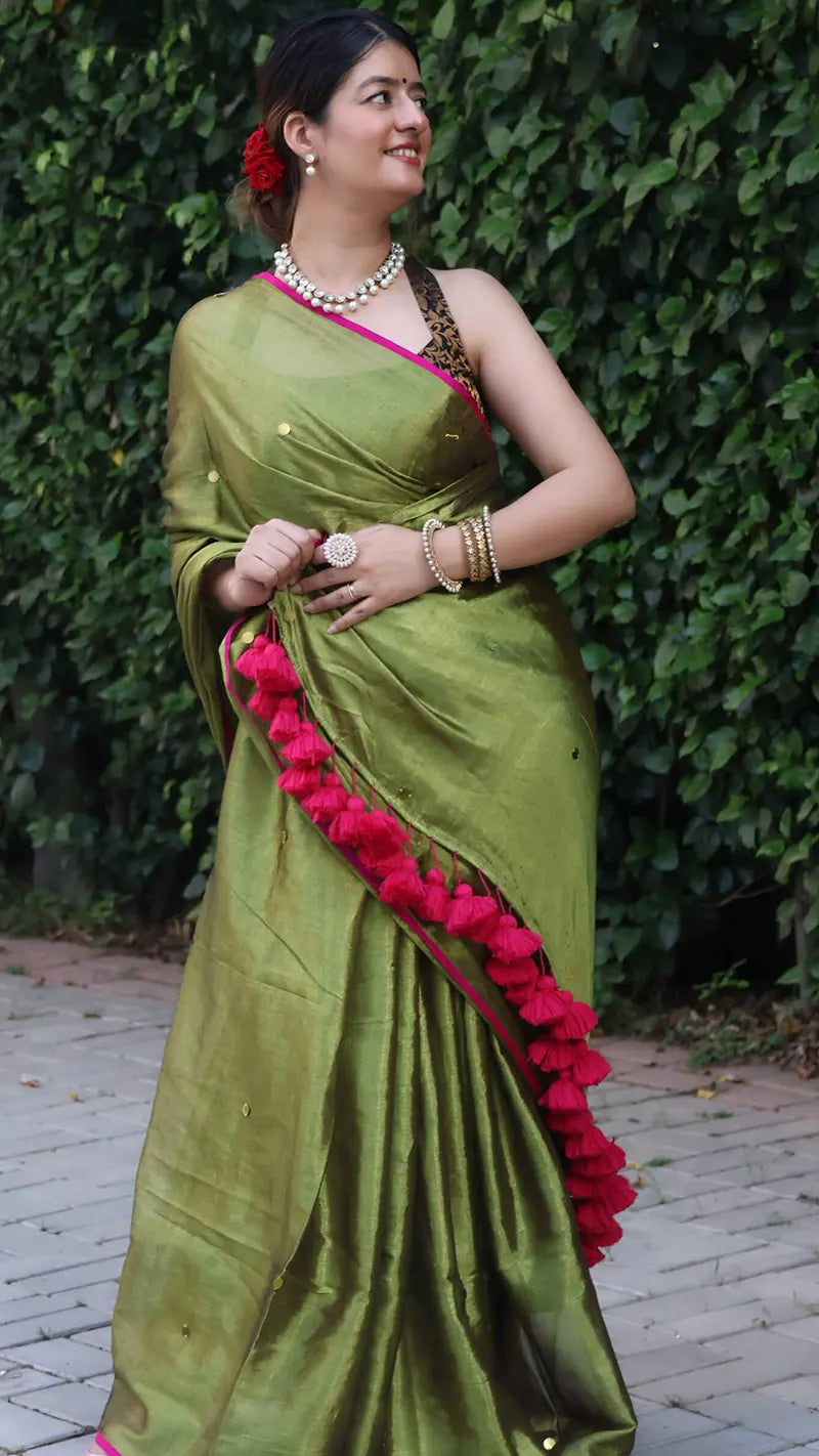 Woman wearing a olive green tissue saree with large ginni chumki against a green leafy background