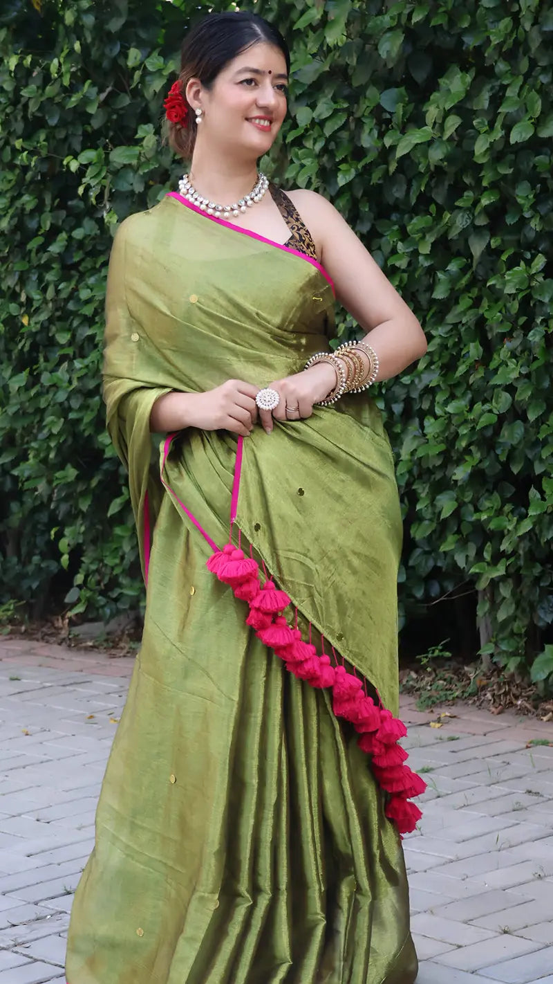 Woman in a olive green tissue saree with large tassels standing against a green leafy background