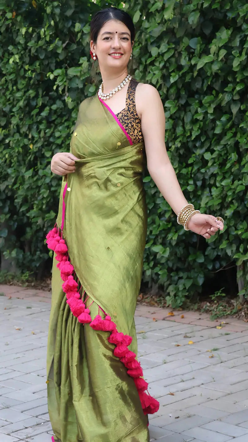 Woman in a olive green tissue saree with large tassels standing against a green leafy background
