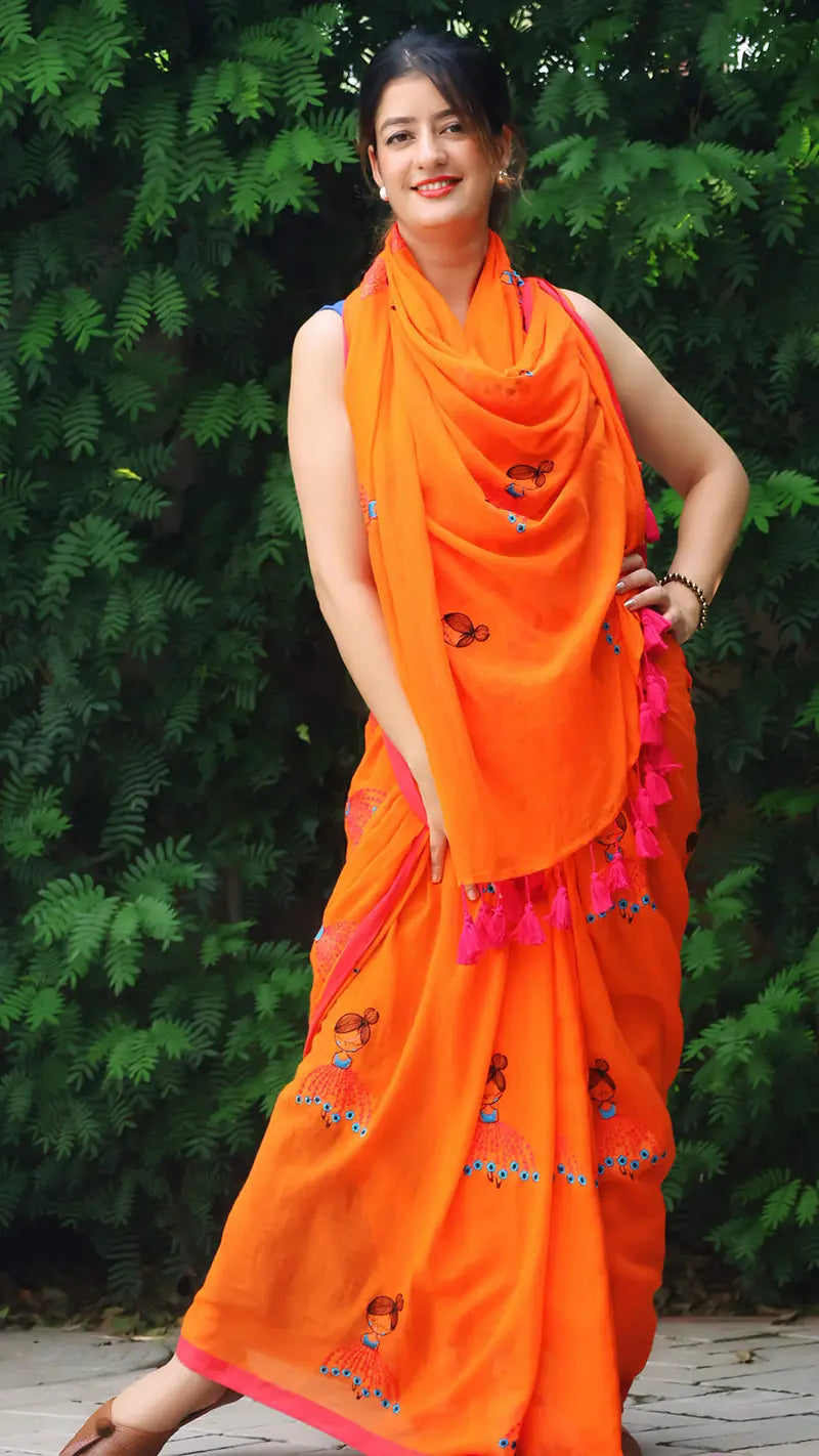 Woman wearing an embroidered orange mul cotton saree with large tassels with a green leafy background