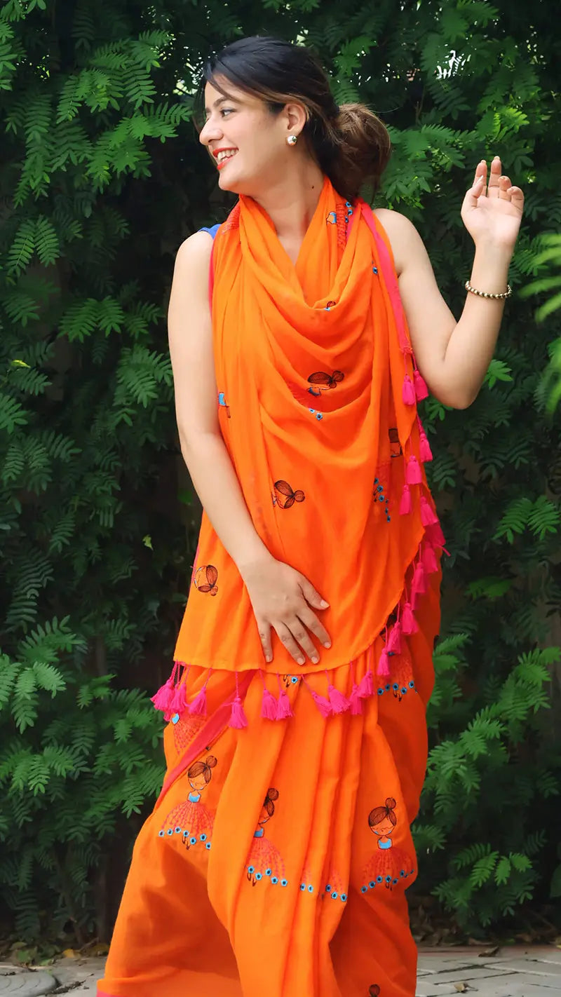 Woman wearing an embroidered orange mul cotton saree with large tassels against a green leafy background