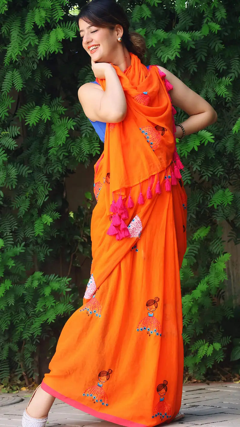 Woman wearing an embroidered orange mul cotton saree with large tassels against a green foliage background