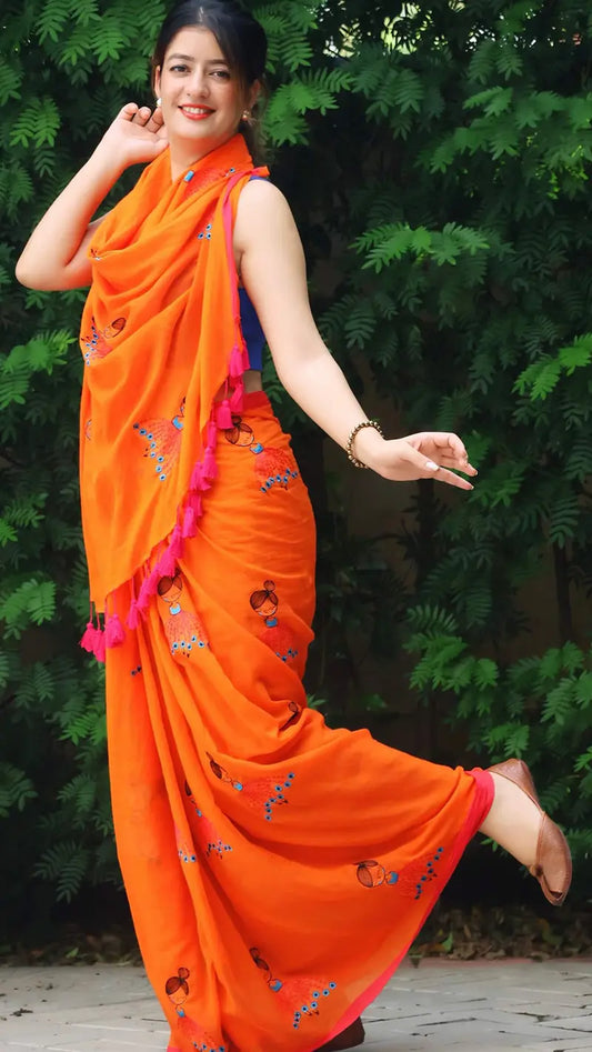 Woman in an orange saree with doll patterns standing against a green leafy background