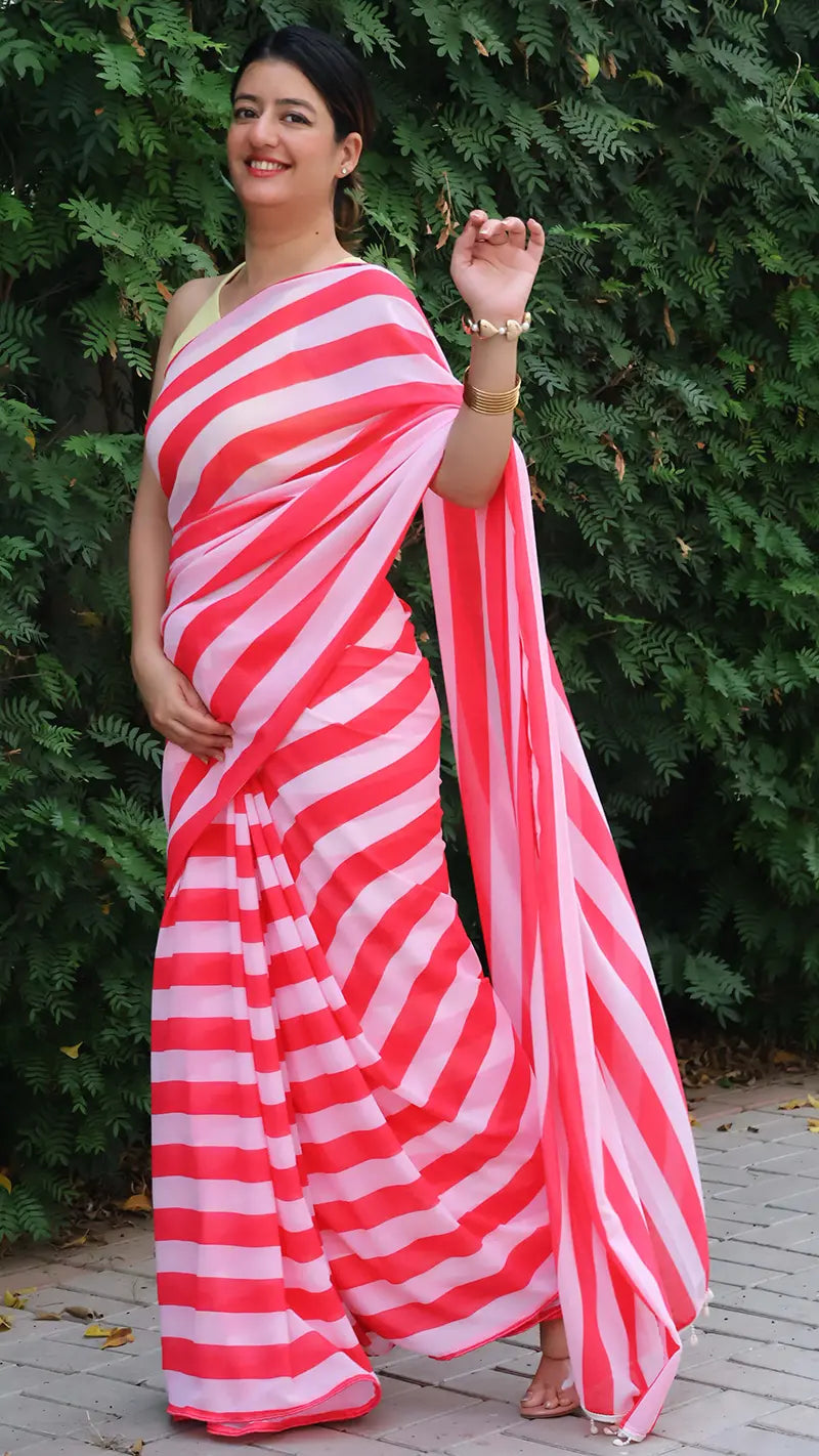 Woman wearing a red and white striped saree standing against a green hedge.