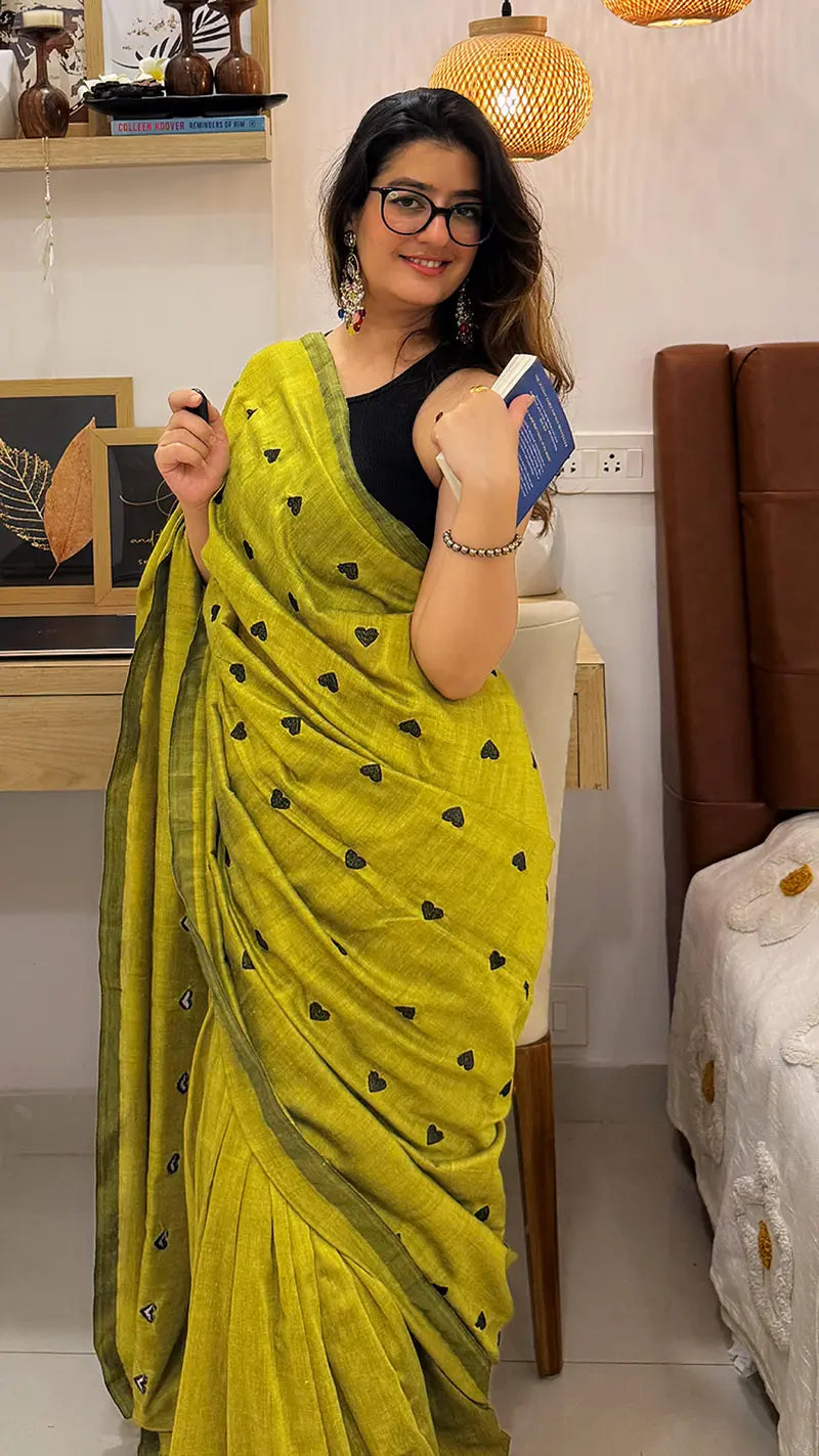Woman wearing a green soft cotton saree with black heart embroidery in a room.