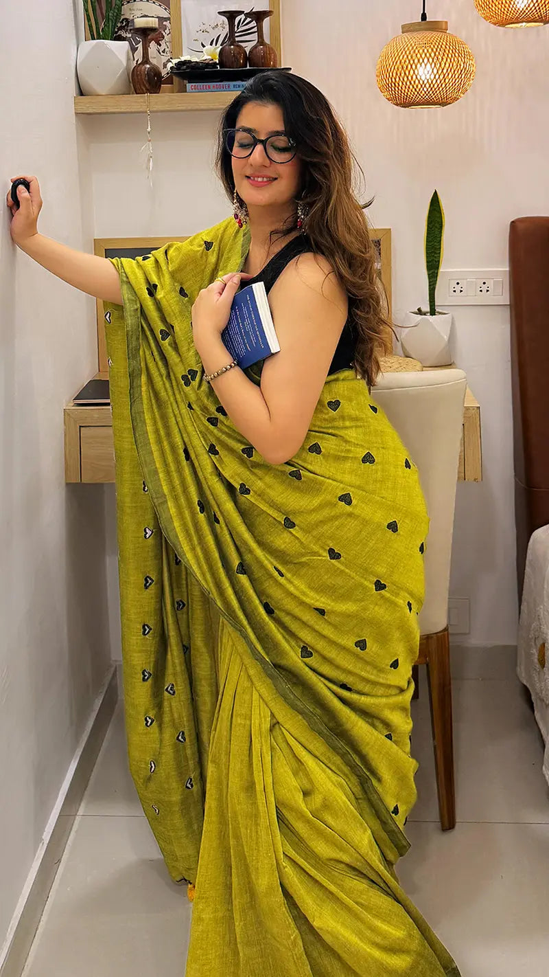 Woman in a green soft cotton saree with black heart embroidery, holding a book, in an indoor setting.