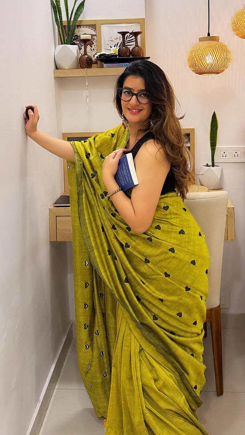 Woman in a green soft cotton saree with black heart embroidery, standing indoors.
