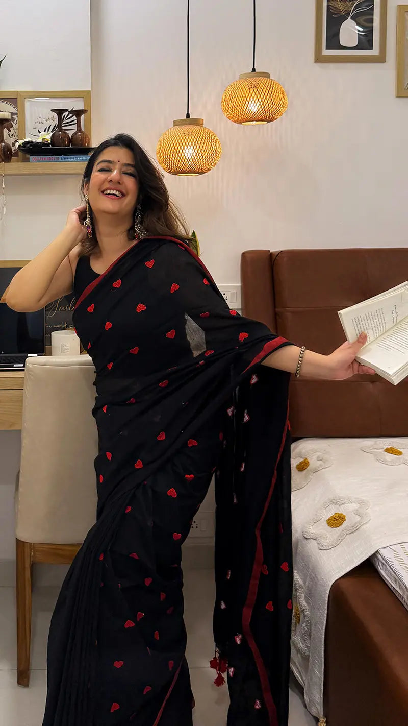 Woman in a black soft saree with red heart embroidery standing in a room with a bed and lampshades.