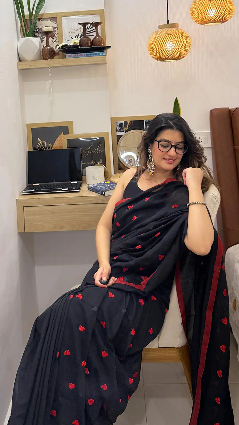Woman in a black soft saree with red heart embroidery sitting in a room with a desk and decor.