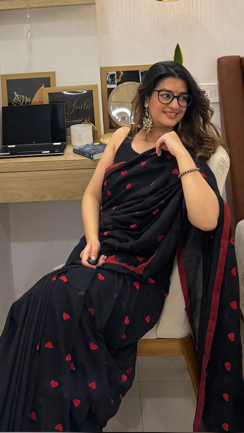 Woman in a black soft cotton saree with red heart embroidery sitting indoors.