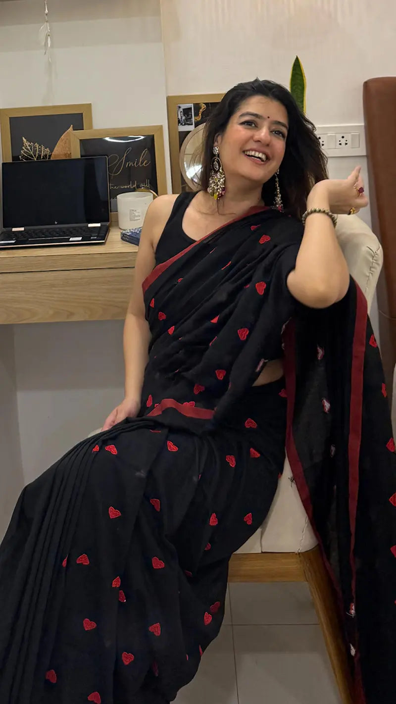 Woman in a black soft saree with red heart embroidery  sitting indoors.