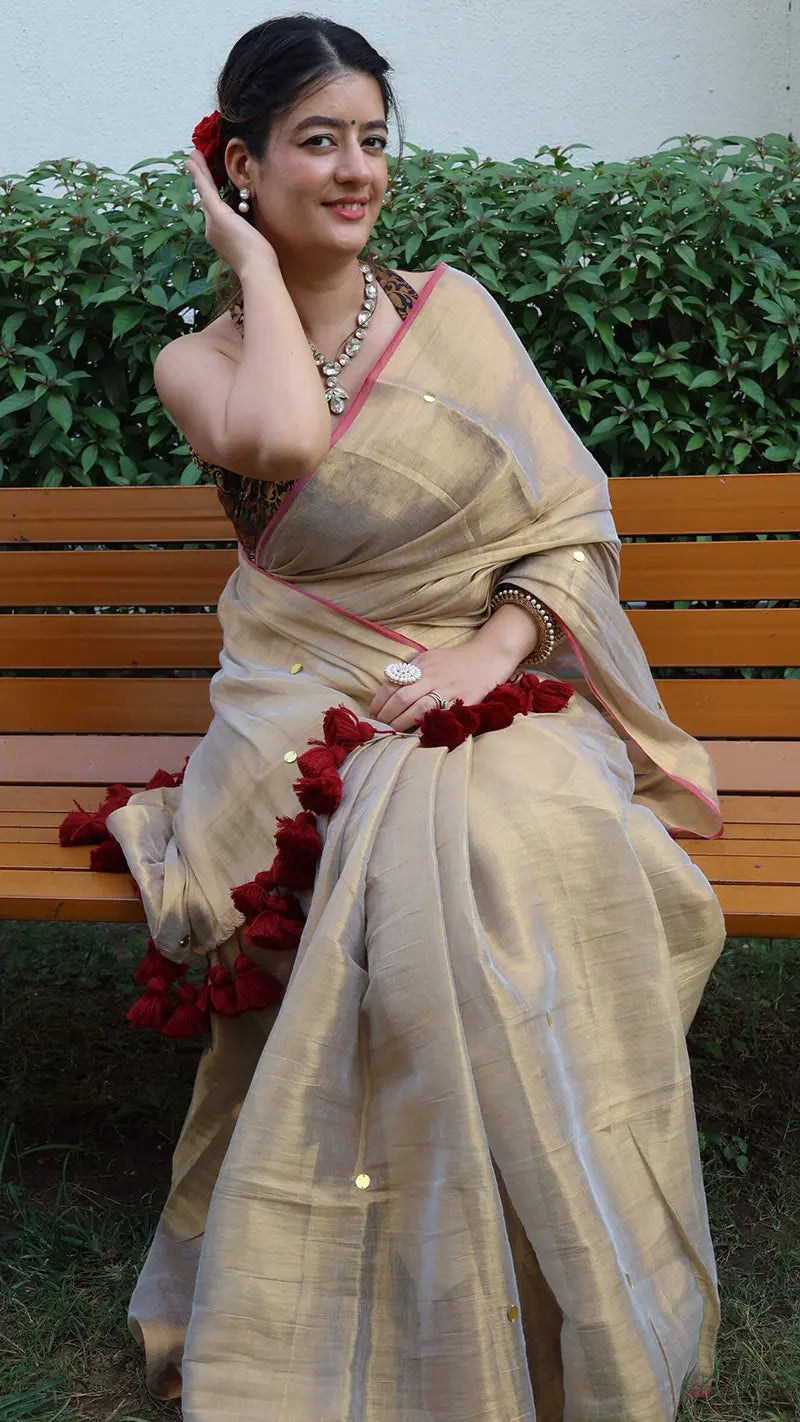 Woman in a hilary gold color tissue saree with red tassels sitting on a bench outdoors.