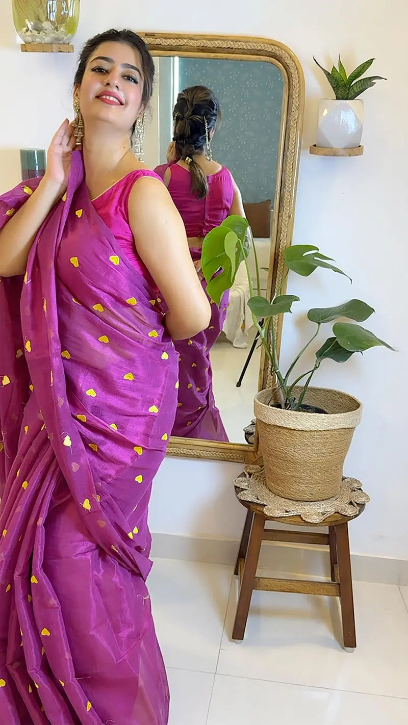 Woman in a pink saree with yellow patterns standing in front of a mirror, with a plant on a small wooden stool beside her.