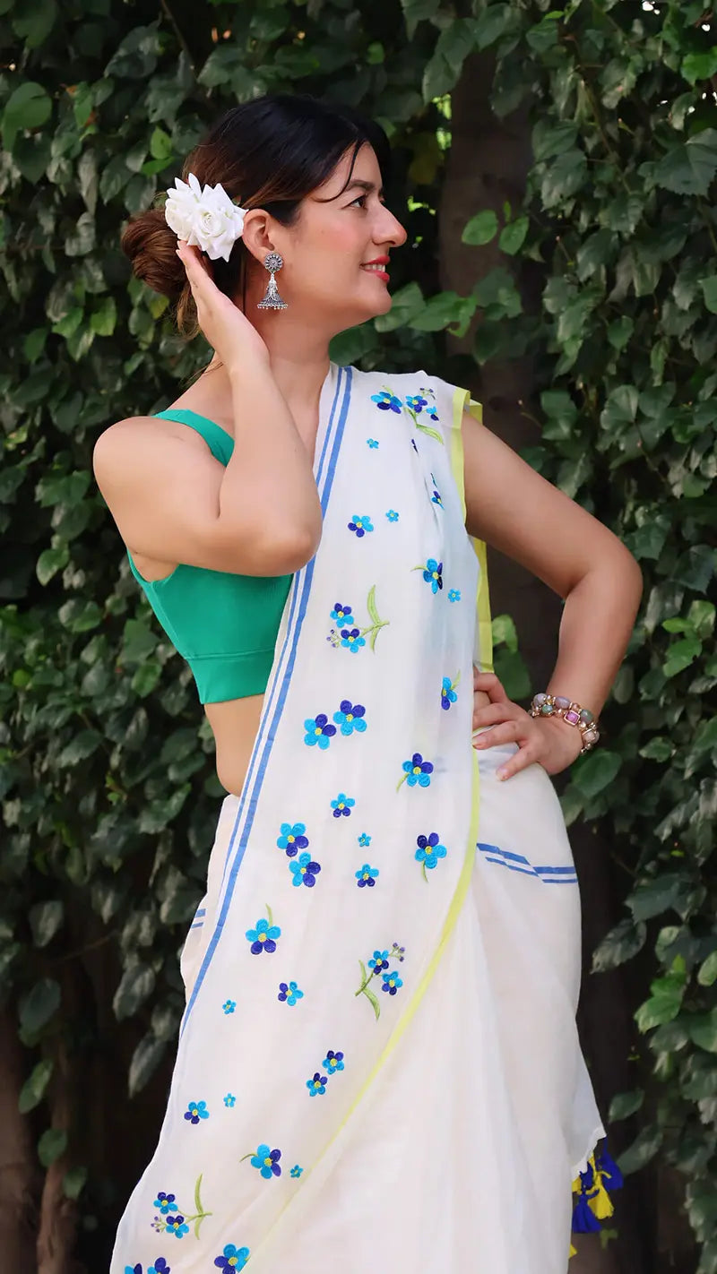 Woman wearing a white mul cotton saree with blue floral patterns against a green leafy background