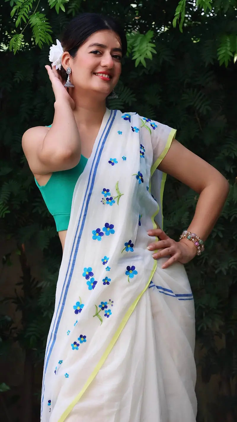 Woman wearing a white saree with blue floral patterns against a green leafy background