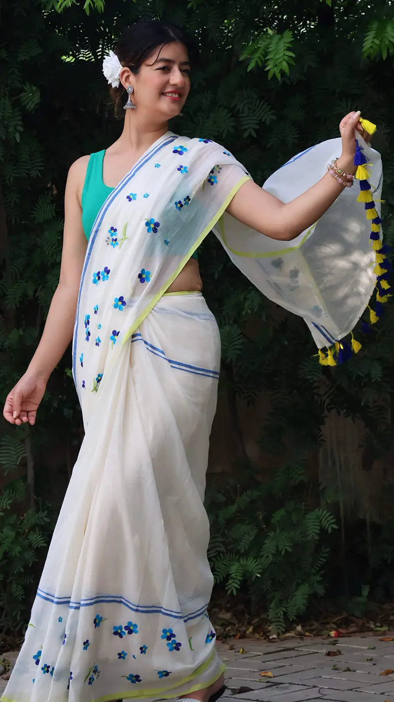 Woman in a white saree with blue floral patterns and a green blouse, standing outdoors.